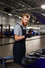 A male car mechanic stands in front of the hood of a car at a service station. An employee checks the oil level in the engine