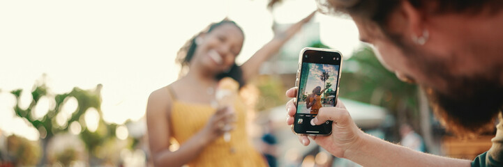 Close-up of a man taking a picture of a woman on a mobile phone. Closeup, smiling woman eating ice cream and filming it with smartphone on urban city background. Backlight