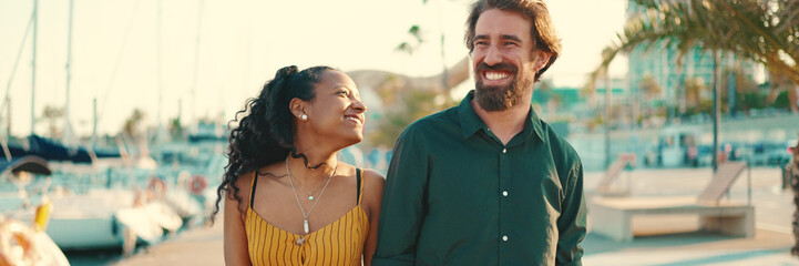 Close-up of young happy man and woman walking along the promenade holding hands and joyfully chatting on yacht background. Closeup, joyful date of a young interracial couple. - obrazy, fototapety, plakaty