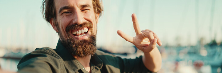 Closeup portrait of a smiling man with a beard chatting on the embankment, on a yacht background. Frontal close-up of happy young male hipster using mobile phone - obrazy, fototapety, plakaty