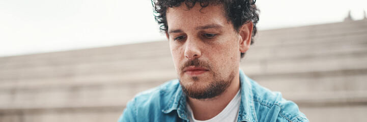 Obraz premium young bearded man in denim shirt sitting on high steps and using mobile phone. Man playing on smartphone
