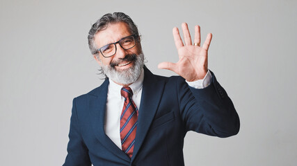 Middle-aged businessman in glasses with gray hair, smiling and waving his hand in greeting, looking at the camera, isolated on gray background in the studio