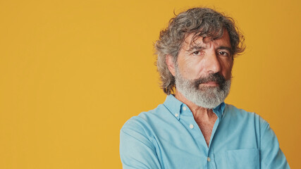 Close-up of an elderly grey-haired bearded man wearing a blue shirt, turning his head and looking frustrated at the camera, isolated on an orange background in the studio