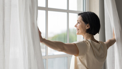 Side view rested middle-aged woman standing near window, open curtains, admires city view, enjoy peaceful, quiet morning, woke up early on weekend, welcoming, start new day, full of hope of happy day - obrazy, fototapety, plakaty