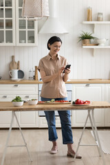 Attractive middle-aged woman use mobile application search recipes on internet, stand alone in cozy domestic kitchen with smartphone in hands, cooking vegetarian salad, share messages. Culinary, tech