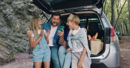 Pretty family mother father and children sitting in car with trunk open laughing browsing online swiping on smartphone. Happy family with their son and daughter using phone sitting in trunk of car