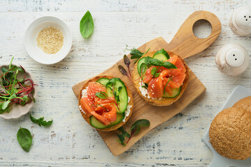 Open smoked salmon sandwiches with cream cheese, cucumber, sesame seeds, microgreens, spinach, and peas leaves on light old wooden background. Healthy breakfast food. Delicious snack. Top view.