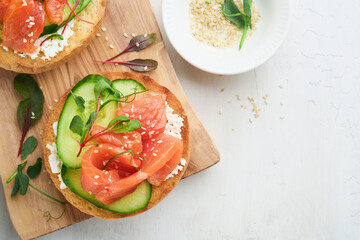 Open smoked salmon sandwiches with cream cheese, cucumber, sesame seeds, microgreens, spinach, and peas leaves on light old wooden background. Healthy breakfast food. Delicious snack. Top view.