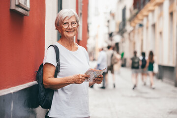Smiling carefree senior woman tourist walking in old town of Seville, Spain holding a backpack consulting a map enjoying vacation trip freedom, healthy lifestyle in retirement