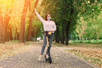 Caucasian female wearing protective face mask riding scooter in city park during covid pandemic.