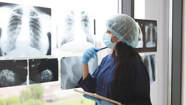 Back view of female surgeon in special clothes looking on patient's chest x-ray attached on window of private clinic. Competent middle-aged doctor using digital tablet for trauma examination.