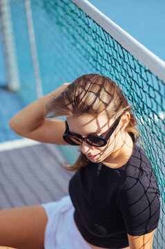 Portrait of a beautiful female tennis player sitting near the net on the tennis court and posing for the camera.