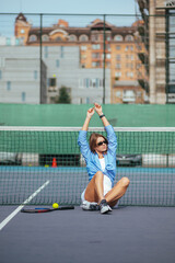 Obraz premium Attractive female tennis player in shirt sitting on court near net with arms raised and posing for camera