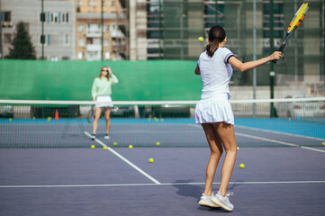 Fototapeta premium The coach trains the student to hit balls. Two women practice playing tennis on the court. Tennis training of two women.