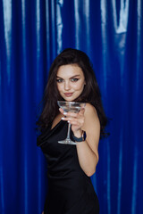 An attractive brunette with a glass of alcohol in her hand stands on a blue background and looks at the camera with a smile on her face.