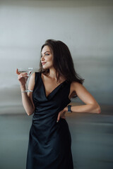 Stylish portrait of an attractive woman in a black dress on a silver background with a glass of...