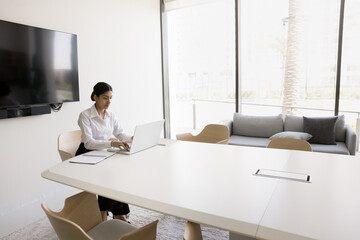 Confident young Indian business professional woman working at laptop alone in meeting room, sitting...