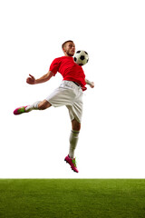 Young man, professional soccer player in red sportwear and boots performing football tricks against white background with green grass.