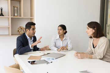 Obraz premium Engaged serious Indian business leader man talking to diverse colleagues women at meeting table, discussing work tasks, management strategy, teamwork. Multiethnic team brainstorming on work ideas
