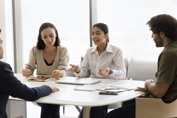 Positive motivated young Indian professional woman offering creative idea for project to listening coworkers on brainstorming meeting, sitting at table, speaking, smiling, working with team