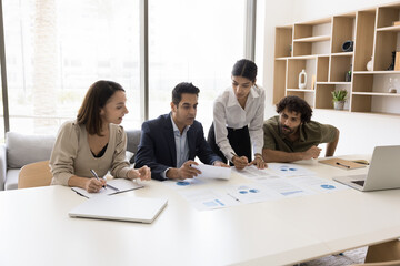 Serious Indian team leader woman presenting sales reports to team. Company coach giving marketing seminar to staff, diverse group of employees, explaining paper diagram on table