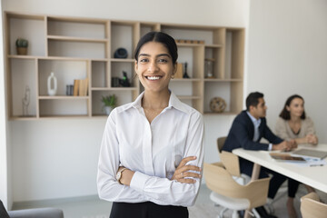 Obraz premium Cheerful manager girl posing with arms folded in office meeting room, looking at camera, smiling, enjoying career, leadership, job in successful company. Confident young business woman portrait