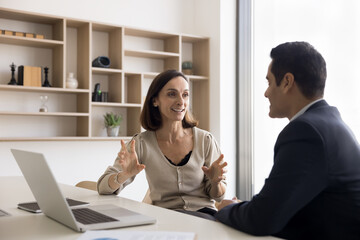 Positive mature business professional woman talking to Indian male colleague at laptop, speaking, smiling, explaining creative idea for project strategy, cooperation, teamwork