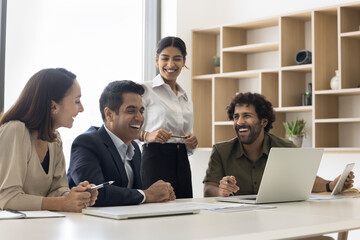 Cheerful Indian and Arab business colleagues laughing at laptop, having fun, relaxing, brainstorming on creative ideas, getting motivation, inspiration, ejoying teamwork spirit, joke, humor