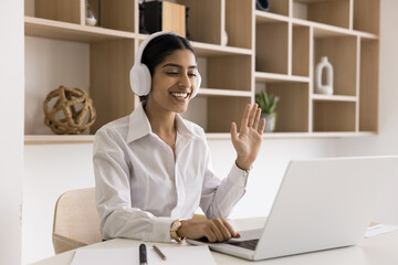 Cheerful young Indian manager woman in headphones online meeting, job communication on Internet, talking on video conference call, waving greeting hand hello at laptop