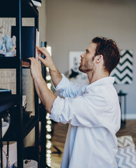 intelligent hipster guy choosing literature on bookcase for autodidact in apartment