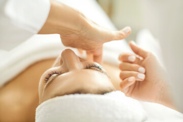 Close up masseur hands making face relaxing massage to a young girl lying with closed eyes in beauty salon or cosmetology cabinet. Professional cosmetologist making massage for woman in spa.