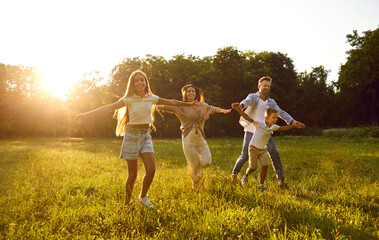 Fototapeta premium Happy family running and having fun in summer park enjoying sunny day in nature. Mother, father and their cheerful two children boy and girl walking on green grass outdoors at sunset together.