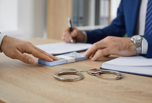 Man hands over cash money to a bondsman asking to release his friend on bail. Unrecognizable businessman gives a bribe to a government official. Crop shot. Handcuffs on a wooden office table close up