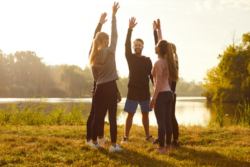 Group of happy people having fitness workout in nature. Team of smiling fit male female athletes standing in circle and raising hands up on grassy bank with misty river in background. Sport concept