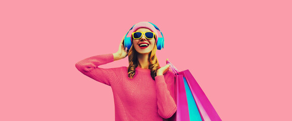 Portrait of stylish happy smiling young woman with colorful shopping bags posing listening to music in headphones wearing knitted sweater, hat on pink studio background