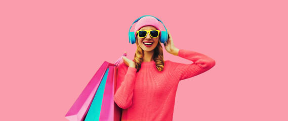 Portrait of stylish happy smiling young woman with colorful shopping bags posing listening to music in headphones wearing knitted sweater, hat on pink studio background - obrazy, fototapety, plakaty