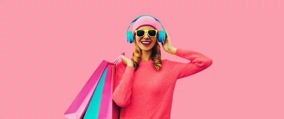 Photo sur Plexiglas Magasin de musique Portrait of stylish happy smiling young woman with colorful shopping bags posing listening to music in headphones wearing knitted sweater, hat on pink studio background