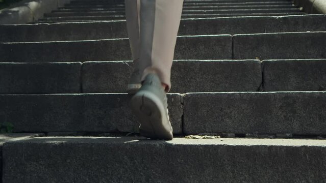 Woman coming up the stairs. Female legs in sneakers going up over stone staircase in city on sunny day. Closeup backside view
