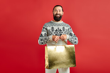 smiling bearded man in sweater with ornament holding shopping bags on red backdrop, Merry Christmas