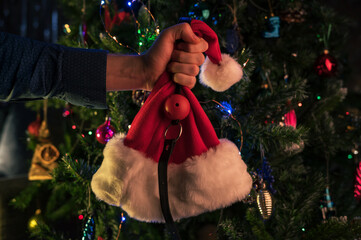Male hand keep BDSM sex toys and santa hat on the christmas tree background