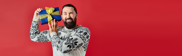 displeased bearded man in winter sweater with ornament holding Christmas present on red, banner