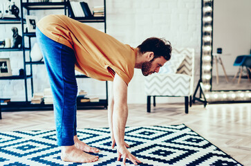 Concentrated young man holding hands on carpet doing morning yoga at home interior