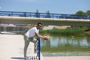 Handsome young man dressed in casual clothes and sunglasses leaning on the railing next to the lake in the park, the man holds a shadow in his hand and enjoys the holiday in seville.