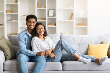 Relaxed indian couple sitting on a couch, embracing and smiling together at camera