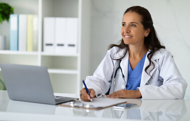 Smiling mature woman doctor taking notes in medical chart - obrazy, fototapety, plakaty