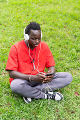 Young man of African ethnicity listening to music with his phone sitting in a park wearing a red shirt.