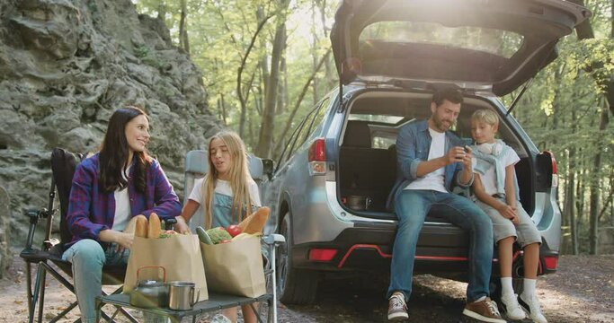 Woman and girl preparing food and father and son siting in open trunk of car and using phone. Happy family having weekend picnic outdoors in the forest. Time to Travel concept. Family travel dream