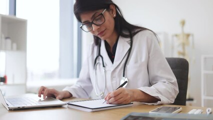 Side view of indian female doctor using portable computer while sitting at office desk in health center interior. Efficient mature general practitioner checking patient records via gadget. - Powered by Adobe