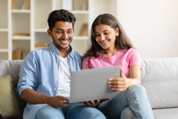Cheerful young Indian couple on couch, sharing a laptop moment