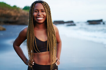 Portrait of positive cheerful sportwoman with dreads standing on beach washed by ocean water